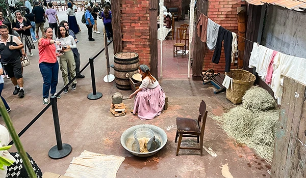 Imagem de uma mulher em traje típico, com atividades tradicionais, cercada por visitantes na Vila Cenográfica da típica Festa da Uva de Jundiaí.