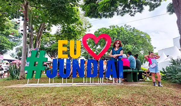 Imagem colorida de um letreiro com a frase "EU AMO JUNDIAI" em um evento ao ar livre, com pessoas se divertindo ao fundo.