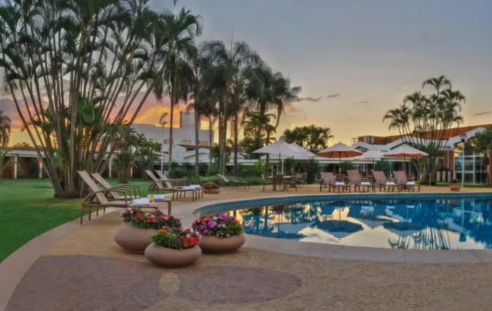 Vista de uma piscina rodeada por palmeiras e flores, ideal para relaxar em um ambiente tranquilo ao pôr do sol. O Royal Tulip JP é perfeito como um dos hotéis para o Carnaval.