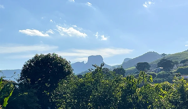 Vista panorâmica de montanhas ao fundo, com céu azul e nuvens, cercada por uma vegetação verde exuberante. Ideal para amantes da natureza e do ecoturismo.