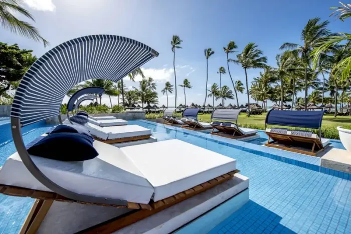 Espaço relaxante à beira da piscina com espreguiçadeiras confortáveis e sombra, cercado por palmeiras sob um céu azul ensolarado.