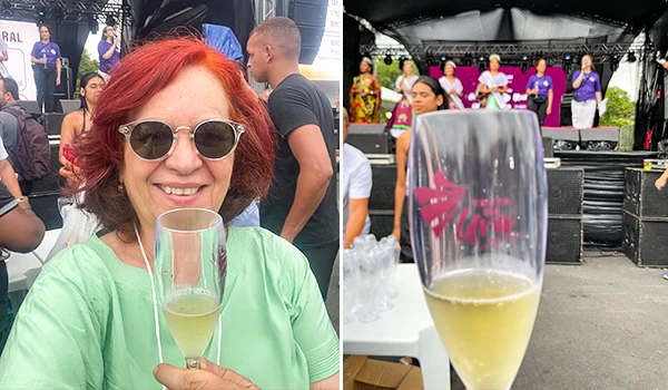 Deusa Rodrigues com cabelo vermelho, usando óculos escuros e blusa verde, segurando taça de espumante em evento ao ar livre com pessoas ao fundo.