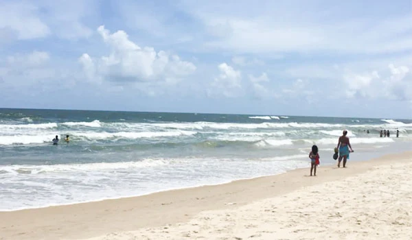 Imagem de uma praia com mar agitado, onde pessoas caminham na areia e crianças brincam na água. Um cenário ideal para férias em família e diversão ao ar livre.