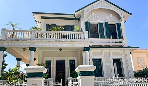 Fachada de uma linda mansão com estilo arquitetônico clássico, apresentando detalhes em branco e verde, cercada por plantas tropicais. Festival do Café em São Paulo.