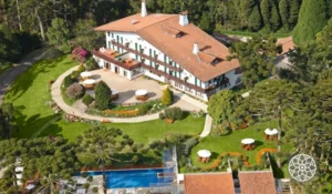 Vista aérea de um elegante hotel com arquitetura charmosa, cercado por um belo jardim e piscina, ideal para relaxar e desfrutar da natureza.