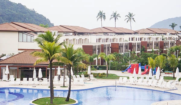 Imagem de um resort com piscina e áreas de lazer, cercado por palmeiras e natureza exuberante, ideal para relaxar em família.