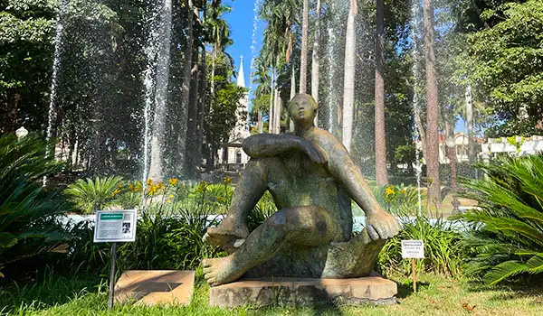 Escultura em bronze de figura humana sentada cercada por natureza e fonte em um espaço público, representando arte e cultura.