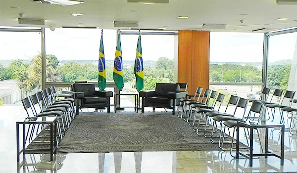 Salão de reuniões com bandeiras do Brasil e cadeiras dispostas para um encontro. Ambiente formal e bem iluminado.