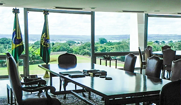Imagem do interior de uma sala do Presidente da República no Palácio do Planalto com vista para a natureza, bandeiras do Brasil ao fundo e mesas de conferência.