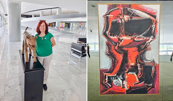 Deusa Rodrigues ao lado de uma escultura de cachorro e um quadro abstrato em um espaço cultural. Destacam-se a arquitetura moderna e as obras de arte.