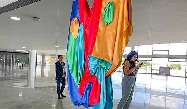 Imagem de uma instalação artística em um espaço moderno, destacando tecidos coloridos pendurados e duas pessoas interagindo com a arte.