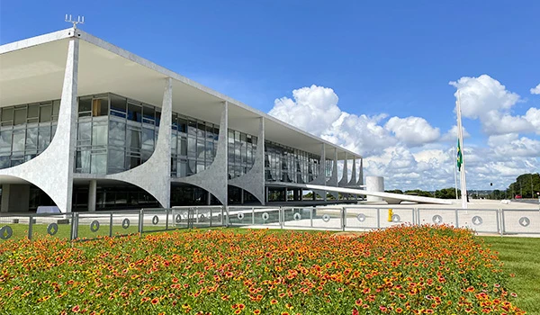 Fachada do Palácio da Alvorada em Brasília, com céu azul e flores ao primeiro plano, representando a arquitetura moderna do Brasil.