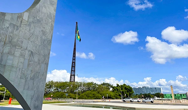 Imagem da bandeira do Brasil tremulando ao vento em destaque, sob um céu azul com nuvens, com um monumento e vegetação ao fundo.
