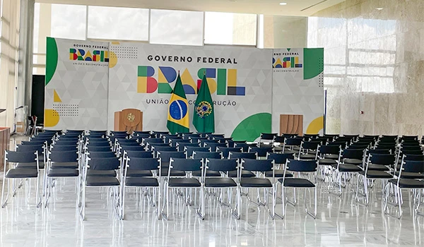 Sala de eventos do Governo Federal do Brasil com cadeiras dispostas para uma reunião. O cenário inclui um banner colorido com elementos da identidade visual do Brasil. Dicas sobre a visita ao Palácio do Planalto.