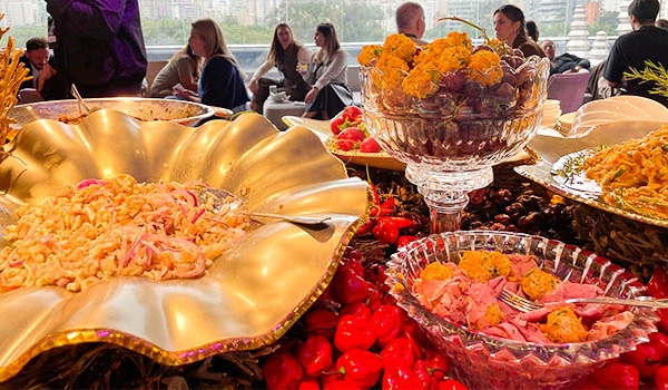 Buffet de comida com destaque para frutas, salgadinhos e pratos coloridos em uma ocasião festiva com pessoas ao fundo. Evento Casar.com para Noivas.