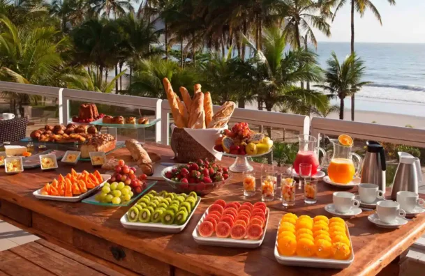 Mesa farta de café da manhã tropical com frutas frescas, pães e bebidas ao ar livre, com vista para o mar e palmeiras ao fundo.