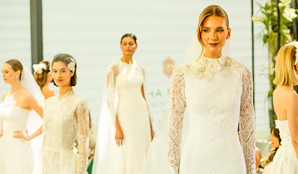 Mulheres vestidas com roupas elegantes, participando de um evento de moda, com foco na mulher ao centro que sorri e veste um vestido de renda branco.