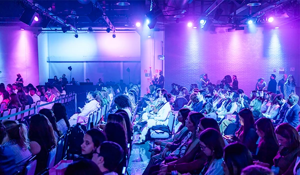 Audiência assistindo a evento com público diversificado em ambiente com iluminação azul e roxa, em sala de conferências ou teatro corporativo.