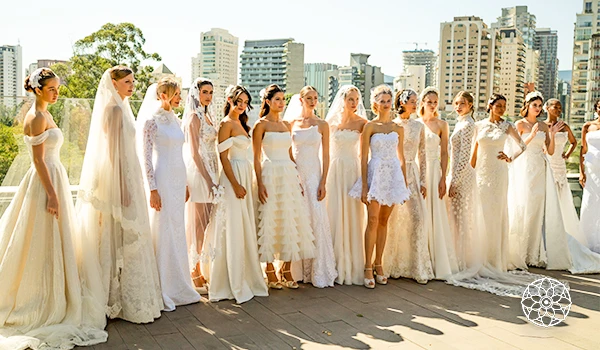 Grupo de noivas vestidas com lindos vestidos de casamento em um evento na cidade, com o céu ao fundo e edifícios modernos ao redor.