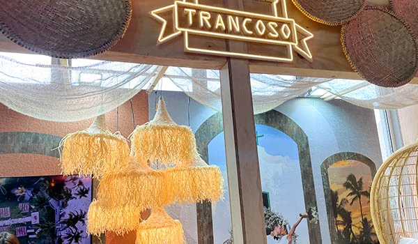 Ambiente decorado com iluminação aconchegante e detalhes sofisticados, incluindo uma luminária de penas e um banner com o nome 'Trancoso', ideal para eventos ou celebrações na atmosfera de Trancoso. Elementos que ressaltam o estilo tropical e elegante.