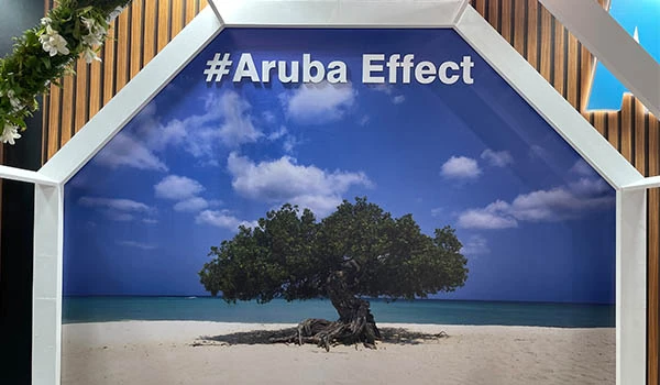 Stand com foto de uma praia de areia branca com uma árvore no centro, sob um céu azul com nuvens, com a hashtag #ArubaEffect no topo, criando um efeito de ilha tropical. Evento Casar.com para Noivas.