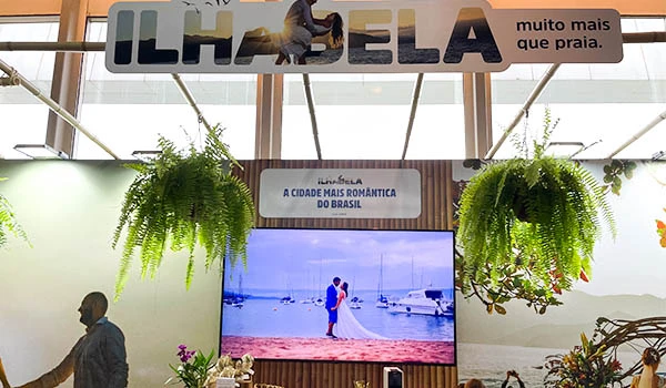 Evento de casamento em Ilha Bela, com vista para o mar, decoração com plantas e televisão exibindo casal na praia, promovendo turismo e romance. Evento Casar.com para Noivas.