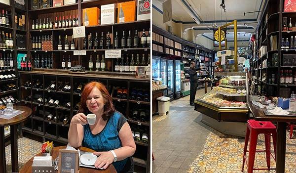 Imagem de Deusa Rodrigues em um café, segurando uma xícara de café, com prateleiras cheias de vinhos e uma vitrine com comidas em um ambiente aconchegante e charmoso. Festival do Café em São Paulo.