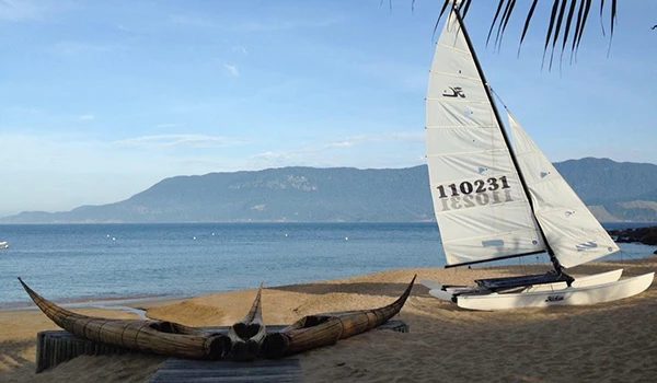 Cena de praia com barco a vela e lenha de canoa tradicional ao pôr do sol, vista para o mar azul e montanhas ao fundo, cenário tranquilo e paradisíaco.