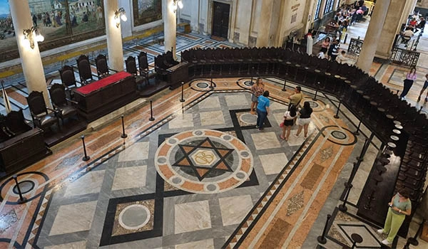 Vista interna de um espaço cultural com detalhes arquitetônicos ricos, incluindo um piso decorativo e disposição de cadeiras.