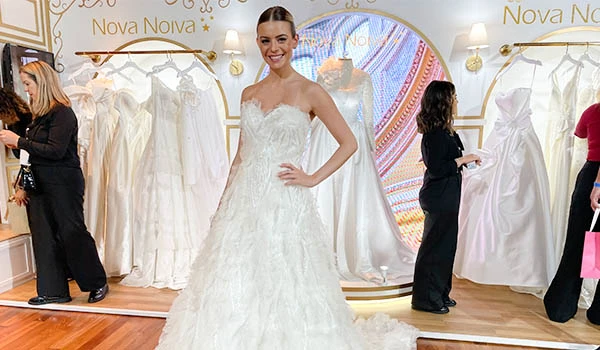 Noiva feliz experimentando vestido de casamento em loja de noivas, com diversas opções de vestidos brancos ao fundo. Evento Casar para Noivas.