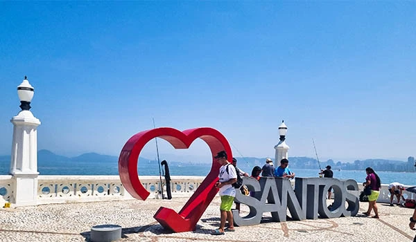 Pessoas em frente ao letreiro "Eu Amo Santos" na orla de Santos, em um dia ensolarado. Uma atração turística famosa na cidade.