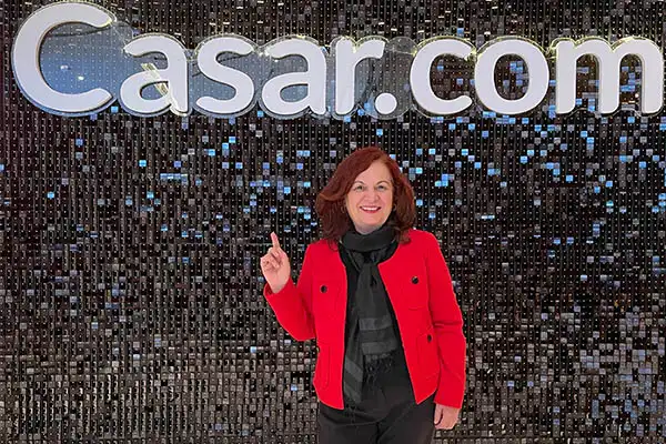 Deusa Rodrigues em evento da Casar.com fundo de luzes decorativas e logo da Casar.com ao fundo, vestida com casaco vermelho e blusa preta.