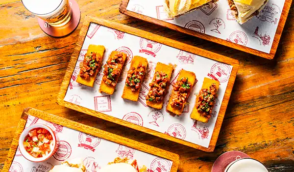 Deliciosos petiscos em uma tábua de madeira, com porções de comida apresentadas de forma atraente, acompanhados de molho e bebida. Ideal para ser um dos presentes para o Dia das Mães com gastronomia.
