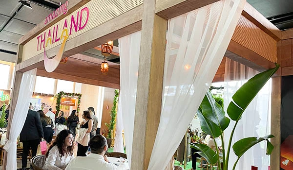 Stand decorado com tema Thailand em evento de turismo, com estrutura de madeira, cortinas brancas, plantas verdes e pessoas conversando ao fundo. Evento Casar.com para Noivas.