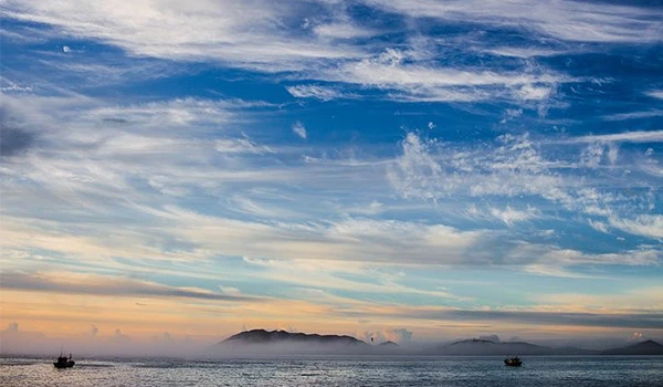 Paisagem marítima ao entardecer com céu azul, nuvens brancas e barcos no horizonte, capturada em um momento de tranquilidade no oceano.