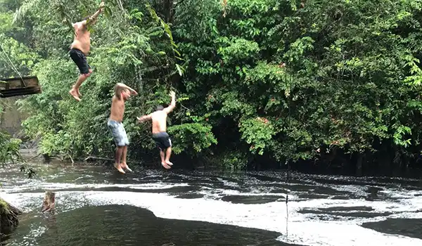 Três meninos pulando de uma ponte de madeira para um rio na natureza, aproveitando um dia de diversão ao ar livre em um ambiente de floresta.
