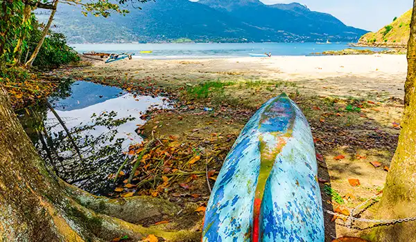 Praia de rio com caiaque à beira d'água e montanhas ao fundo, cenário de natureza com árvores, areia e sereno.