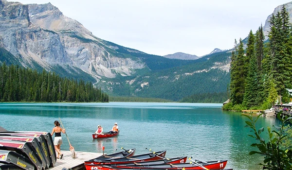 Lago tranquilo cercado por montanhas e árvores verdes, com pessoas preparando caiaques para um passeio em um dia ensolarado na natureza.