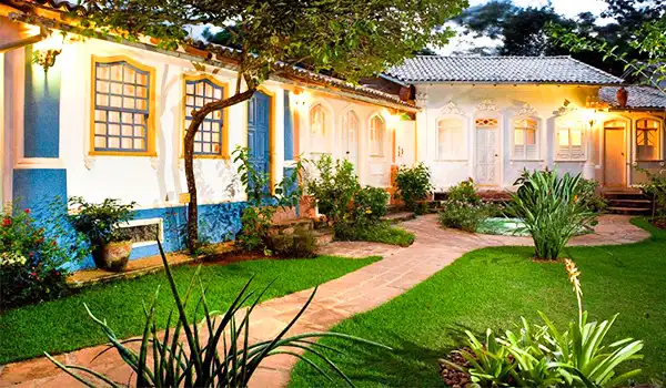 Imagem de uma charmosa casa colonial colorida, com jardim bem cuidado na frente, árvores e iluminação suave, criando um ambiente aconchegante e convidativo.