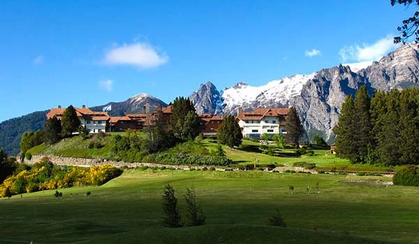 Paisagem de um campo de golfe cercado por árvores e casas, com uma cadeia de montanhas ao fundo sob céu azul claro com algumas nuvens.