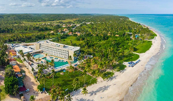 Vista aérea de um resort na praia com piscinas, cercado por vegetação tropical, ao lado do mar azul e areia branca, em um cenário paradisíaco.