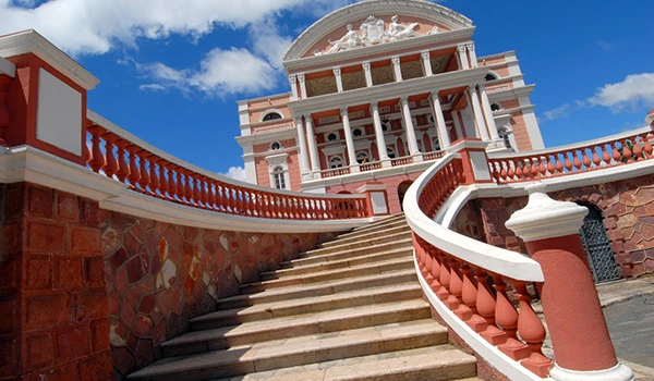 Imagem de um palácio com escadaria e arquitetura clássica, com detalhes em branco e rosa, sob céu azul com nuvens, representando um ponto turístico.