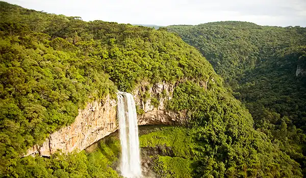 Paisagem com uma cachoeira no meio de uma floresta densa, rodeada por morros verdes e céu parcialmente nublado, destaque para a beleza natural e tranquilidade do local.