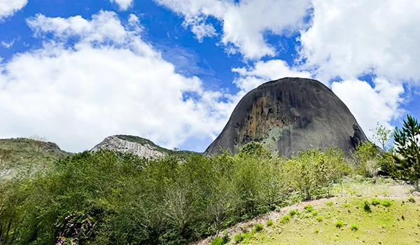 Imagem de uma formação rochosa icônica na natureza com árvores verdes ao redor, céu azul com nuvens brancas. Férias na Montanha ou na Praia.