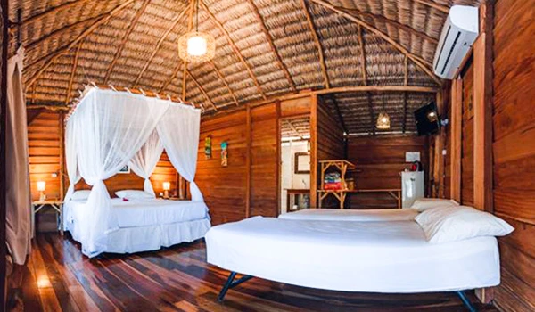 Interior de um bangalô de madeira com móveis simples, cama de casal com dossel, decorado com cortinas brancas e iluminação aconchegante, ideal para hospedagem confortável na natureza.