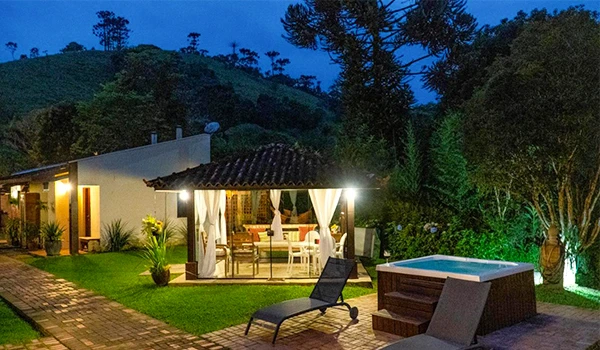 Imagem noturna de uma residência ao ar livre com área de lazer, incluindo spa, espreguiçadeiras e uma área coberta com móveis confortáveis, cercada por natureza exuberante.