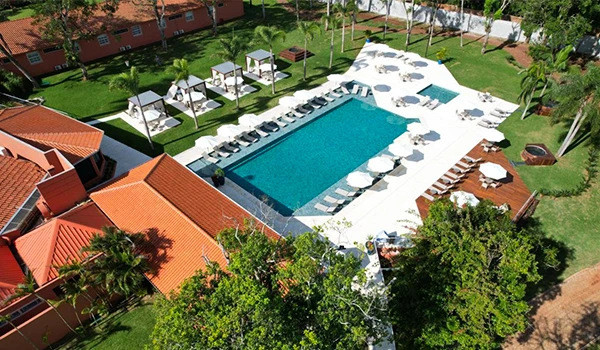Imagem aérea de uma piscina grande e moderna em um resort ou hotel, rodeada por espreguiçadeiras, cabanas, áreas verdes e estruturas com telhados de barro.