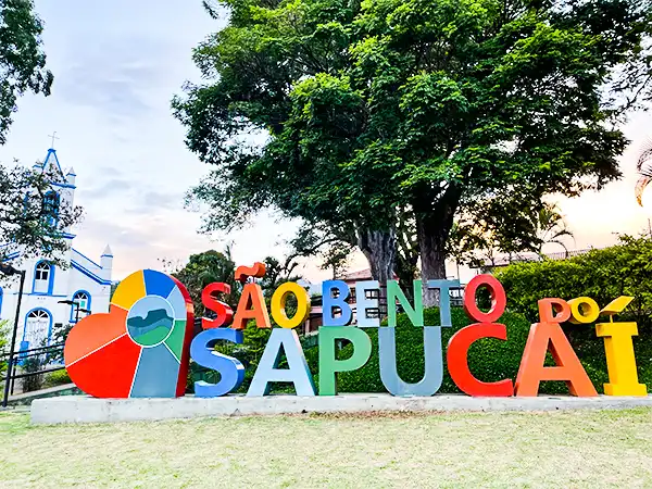 Letreiro turístico de São Bento do Sapucaí com cores vibrantes e uma grande árvore ao fundo, ideal para fotos e visitar pontos turísticos na cidade.