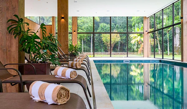 Piscina interior com cadeiras de descanso e toalhas, ambiente elegante e bem iluminado com vista para área verde externa, ideal para relax.