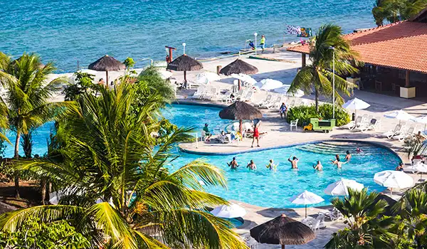 Piscina de resort tropical com coqueiros, espreguiçadeiras e vista para o mar, ideal para férias e lazer ao ar livre.
